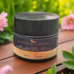 Jar of Luneria Beauty Botanica product on a wooden surface with flowers in the background