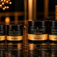body care products displayed on a dark reflective surface with warm decorative lighting. The set includes two smaller jars labeled Trial Size and two larger jars labeled Starter Size, featuring black lids and gold-and-black labels.